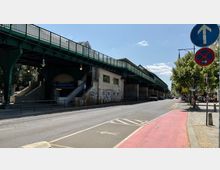 Straßenszene mit einer erhöhten, grünen U-Bahn-Brücke und dem Eingang zur Station "Eberswalder Straße" darunter. Rechts verläuft eine Straße mit Radweg und Gehweg, flankiert von Bäumen und Verkehrsschildern.