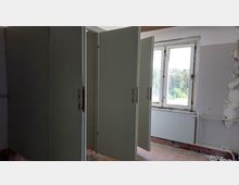 Ein verlassener Innenraum mit alten, offenen Metalltüren, Fenster mit Blick auf Bäume im Hintergrund und abblätternder Wandfarbe. Am unteren Rand ist ein roher Ziegelboden und ein Heizkörper unterhalb des Fensters sichtbar.