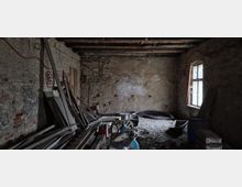 Renovierungsbedürftiger Raum mit unverputzten Backstein- und Putzwänden, freiliegenden Holzdeckenbalken und einem Fenster, das Tageslicht hereinlässt. Im Raum befinden sich Baumaterialien, Werkzeuge und verschiedene Gegenstände wie Plastikstühle und Fässer.