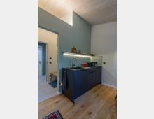 Moderne, kleine Küchenzeile mit dunklem Unterschrank, eingebauter Spüle und einer dezenten Unterbeleuchtung an einer mattgrünen Wand. Der Boden besteht aus hellem Holz, die Decke ist aus unbehandeltem Holz, und im Hintergrund ist eine Tür zu einem angrenzenden Raum sichtbar.