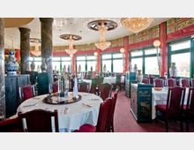 Ein elegantes chinesisches Restaurant mit runden, weiß gedeckten Tischen, roten Holzstühlen und dekorativen Elementen wie Porzellanvasen, Marmorsäulen und kunstvollen Kronleuchtern. Große Fenster an einer Wand lassen viel Tageslicht in den Raum.