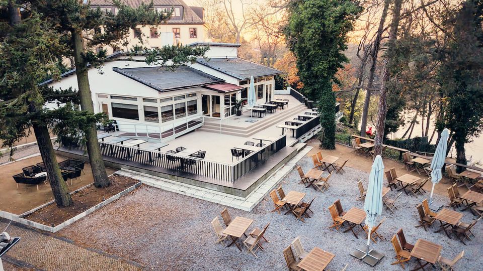 Elevated view of a modern, low white building with large windows and a fenced terrace, surrounded by tall trees. In the foreground is a gravel outdoor seating area with wooden tables and chairs and several closed patio umbrellas near a waterfront.