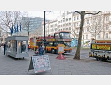 Straßenszene mit einem offenen Doppeldeckerbus für Stadtrundfahrten, beschriftet mit „Sightseeing“, geparkt an einer Haltestelle neben einem Gehweg. Im Vordergrund befinden sich ein Informationsstand, Werbetafeln mit Stadtrundfahrt-Angeboten und Menschen, die Tickets kaufen. Im Hintergrund sind Gebäude mit klassischer und moderner Architektur sowie Bäume ohne Laub zu sehen.