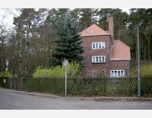 Backsteinhaus mit roten Ziegeldach in ländlicher Umgebung, umgeben von hohen Bäumen und niedrigen Sträuchern. Ein Holzzaun und ein Verkehrsschild trennen das Grundstück vom Gehweg einer ruhigen Straße.