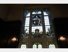 Großes Buntglasfenster in einem historischen Innenraum, verziert mit detaillierten religiösen und ornamentalen Motiven. Das Fenster ist in mehrere Segmente unterteilt, darunter eine zentrale Darstellung und komplexe Rankenmuster; die Umgebung ist durch dunkles Holz und Wandleuchter geprägt.