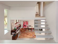 Split-level modern home interior with white walls and a floating gray staircase leading to an upper landing. Below, a small reading or study nook features built-in wood shelving filled with books, framed artwork, and minimalist chairs beside a window.