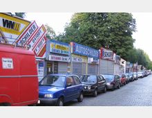 Straße mit mehreren Geschäften, die Schilder- und Versicherungsdienstleistungen anbieten; im Vordergrund parken Autos entlang einer Pflasterstraße, umgeben von Bäumen im Hintergrund.