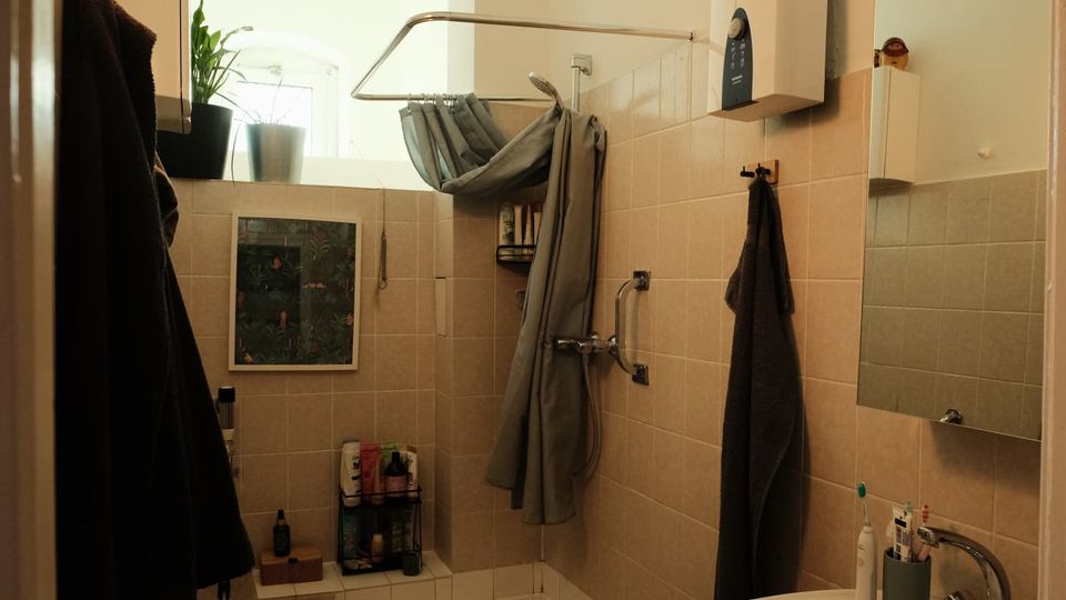 Kleines Badezimmer mit hellbeigen Fliesen, Badewanne mit Duschvorhang und Haltegriff sowie Waschbecken mit Spiegel und Ablage. Ein Fenster mit Pflanzen bringt Tageslicht in den Raum; Handtücher und Badutensilien sind an Haken und Regalen verstaut.
