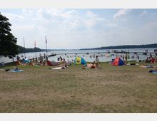 Strandbereich an einem See mit zahlreichen Badenden im Wasser und auf der Wiese. Im Vordergrund liegen bunte Decken und kleine Strandzelte, während im Hintergrund ein langer Steg in den See hineinragt, gesäumt von Badegästen.