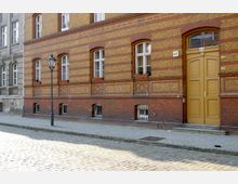 Straßenansicht eines Altbaus mit einer Fassade aus gelb- und rot-braunen Ziegeln, mehreren Fenstern mit Blumenkästen sowie einer gelben Eingangstür. Vor dem Gebäude verläuft eine gepflasterte Straße, daneben ein Gehweg mit einem schwarzen Laternenpfahl.