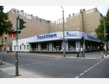 Straßenecke in einer städtischen Umgebung mit einem markanten Geschäft für Textilien und Haushaltswaren, das ein Eckgebäude einnimmt. Die Fassade ist mit großen Schildern beschriftet, und dahinter ragen beige Wohngebäude mit Graffiti auf. Verkehrsschilder und eine Ampel sind im Vordergrund sichtbar.