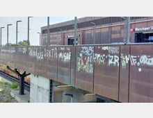 Außenansicht des Bahnhofs Warschauer Straße in Berlin mit einer braunen Metallfassade, die mit verschiedenen Graffiti und Schriftzügen bedeckt ist; im unteren Bereich ist ein Zug der S-Bahn zu sehen.