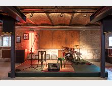 Eine kleine Bühne in einem Backsteinraum mit gewölbter Decke. Auf der Bühne stehen ein Schlagzeug, Mikrofonständer, zwei Stühle und Verstärker, umgeben von Teppichen und roten Neonlichtern an der Backsteinwand.