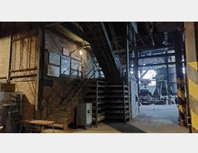 Innenansicht einer alten Fabrikhalle mit Backsteinwänden und Metalltreppe. Im Vordergrund befinden sich Regale, Schilder und eine Werkbank; im Hintergrund sind große metallische Maschinen und offene Arbeitsbereiche sichtbar.