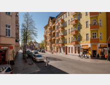 Straßenszene in einer Wohngegend mit mehrstöckigen Altbauten in gelber und orangefarbener Fassade, Balkonen und Geschäften im Erdgeschoss, darunter eine Bäckerei. Im Vordergrund führen ein Bürgersteig, parkende Autos und ein Fußgänger über eine Kreuzung, während Bäume entlang der Straße Schatten spenden.