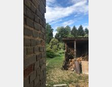 Eine ländliche Szene mit einer gemauerten Hauswand links im Bild, einem Schuppen mit gestapeltem Brennholz rechts und einem Garten mit Bäumen und Wiese im Hintergrund. Der Himmel ist teils bewölkt, und im Vordergrund steht eine Schubkarre vor dem Schuppen.