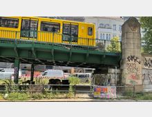 Ein gelber Zug der Berliner U-Bahn fährt über eine grüne Stahlbrücke, die von massiven steinernen Pfeilern mit Graffiti getragen wird. Unter der Brücke befinden sich überwucherte Pflanzen, Möbelreste und Fahrzeuge, während im Hintergrund Gebäude und Baustellenabsperrungen zu sehen sind.