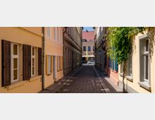 Schmale, gepflasterte Gasse mit historischen Gebäuden in Gelb- und Beigefarbtönen, teils mit Fensterläden und einem Baum auf der rechten Seite. Im Hintergrund ein weiteres Gebäude mit roten Dachziegeln und geparkten Autos.