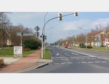 Breite Stadtstraße mit mehreren Fahrspuren und Markierungen, flankiert von Fuß- und Radwegen sowie grünem Rasenstreifen mit Bäumen. Im Hintergrund befinden sich Wohngebäude mit orangefarbenen Dächern und ein Kirchturm.