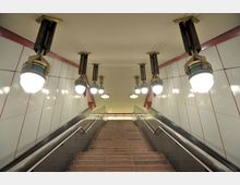 Treppe in einer U-Bahn-Station mit symmetrisch angeordneten Hängelampen im Retro-Design. Die Wände sind mit weißen Kacheln und roten Linien gestaltet, und seitlich befinden sich Handläufe aus Metall.