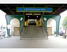 Eingang zum U-Bahnhof Schönhauser Allee in Berlin unter einer grünen Metallbrücke, mit einer breiten Treppe, die zum Bahnhof hinaufführt. Links und rechts sind geschwungene Arkaden sowie Fahrräder und parkende Autos sichtbar.