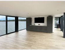 Modernes Interieur eines schwimmenden Hauses oder Gebäudes mit großen bodentiefen Fenstern, die auf eine Wasserlandschaft blicken. Im Raum befindet sich eine dunkelgraue Küchenzeile mit integriertem Waschbecken und flachem Bildschirm; der Boden besteht aus hellem Holz.