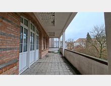 Langer, überdachter Balkon oder Laubengang an einem Backsteingebäude mit weißen Fenstern und Türen, Fliesenboden und Betondecke mit Lochmuster. Rechts verläuft eine niedrige Brüstung mit Blick auf Bäume und umliegende Wohnhäuser.