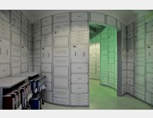 Ein Raum mit Wänden, die mit Paneelen aus einer Vielzahl von Schaltern, Knöpfen und Anzeigen bedeckt sind, ähnlich einer technischen Schalttafel. Ein gekrümmter Durchgang führt in einen hinteren Bereich mit grüner Beleuchtung, und am Boden steht eine Reihe von Koffern oder Containerboxen.