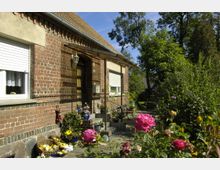 Backsteinhaus mit weißen Fenstern und einem kleinen Vordach über der Haustür, umgeben von einem blühenden Garten mit Rosen und anderen Pflanzen. Im Vordergrund stehen Körbe mit Obst und Gemüse auf einer Terrasse, während Bäume im Hintergrund einen ländlichen Charakter vermitteln.