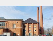 Ziegelgebäude eines ehemaligen Industriekomplexes mit hohen, roten Schornsteinen und rechteckigen Fenstern. Im Vordergrund ist ein Parkplatz, im Hintergrund strahlt ein bewölkter Himmel.