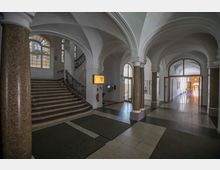 Eingangsbereich eines historischen Gebäudes mit Gewölbedecken, Marmorsäulen und einer Treppe mit kunstvoll verziertem Geländer. Im Hintergrund führt ein heller Korridor mit Glastüren weiter in das Gebäude.