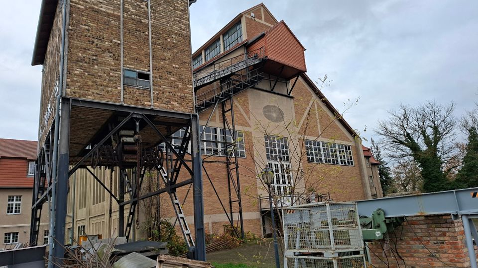 Backstein-Gebäude eines ehemaligen Industriekomplexes mit großen Fenstern, schrägem Dach und außen angebrachten Metalltreppen. Im Vordergrund befinden sich Metallkonstruktionen und Gerüste, im Hintergrund kahle Bäume und ein bewölkter Himmel.