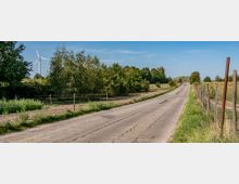 Eine ländliche gepflasterte Straße, flankiert von Zäunen und Wiesen, führt geradeaus in eine von Bäumen gesäumte Landschaft. Im Hintergrund ist ein Windrad hinter Bäumen sichtbar, unter einem klaren blauen Himmel.