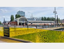 Gelbes Geländer mit Smiley-Motiven und der Aufschrift „East Side Mall“ im Vordergrund, dahinter das Eingangsgebäude des U-Bahnhofs Schlesisches Tor mit einer Glasfassade und umliegender Bebauung; eine Brücke mit Fußgängern und Autos verbindet die Szenerie.