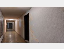 Ein langgezogener, schmaler Flur mit hellem Deckenlicht, abgenutztem Boden und teils beschädigten Wänden. Auf der rechten Seite befinden sich dunkle Türen, links sind Wandflächen mit einer dekorativen Struktur erkennbar.