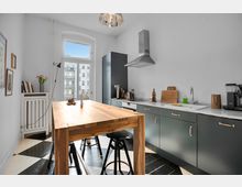 Moderne Küche mit schwarz-weißen Fliesen, dunkelgrünen Unterschränken mit goldenen Griffen und einer langen Arbeitsfläche aus weißem Material. Ein Massivholztisch mit Barhockern steht in der Raummitte, vor einer großen Fensterfront, die Blick auf andere Gebäude bietet.