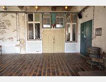 Industrie-innenraum mit rustikalem Holzboden, einer cremefarbenen Doppeltür mit Fenstern darüber und einem Hirschgeweih an der Wand. Die Wände bestehen teils aus weiß gestrichenen Backsteinen und teils aus Putz, ergänzt durch Fenster mit Holzrahmen und industriellen Details wie freiliegenden Kabeln.