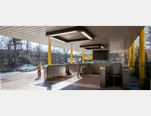 U-Bahn-Eingang mit zwei Rolltreppen, gelben Säulen und einer weißen Decke mit rechteckigen Leuchtelementen; im Hintergrund Bäume und eine Straße mit vorbeifahrendem Auto.