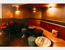 Gemütlicher Bar- oder Lounge-Innenraum mit langen, gepolsterten Sitzbänken aus rotem Kunstleder entlang der Wand, mehreren kleinen runden Tischen und Hockern. Warme Wandlampen und dunkel geflieste Böden prägen die gedämpfte Beleuchtung und das intime Ambiente.