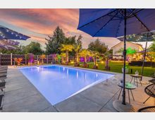 Außenbereich mit einem beleuchteten Swimmingpool, umgeben von Palmen, farbigen Lichtern und modernen Gartenelementen. Im Hintergrund ein Haus, Liegen, Sonnenschirme und gemütliche Sitzbereiche.