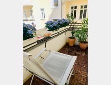 Kleiner Balkon mit Holzfliesen, weißer Liege und Geländer, auf dem mehrere Blumenkästen mit blauen Hortensien stehen. Im Hintergrund sind helle Hausfassaden mit Fenstern sowie zwei große Pflanzkübel mit Sträuchern zu sehen.