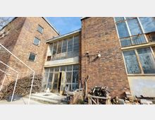 Backsteinfassade eines mehrstöckigen Gebäudes mit großen, teils verglasten Fenstern und Metallgittern an einigen Eingängen und Fenstern. Im Vordergrund sind Holzstapel, Baumäste und Baustellenmaterialien zu sehen.