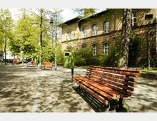 Ein kleiner Park mit roten Holzbänken, grünen Abfallbehältern und gepflasterten Wegen, umgeben von Bäumen; im Hintergrund ein historisches Backsteingebäude mit weißen Fensterrahmen.