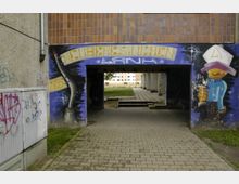 Ein gepflasterter Durchgang unter einem Wohngebäude, dessen Wände mit bunten Graffitis, darunter ein Schriftzug „Neuleresinchen“, bemalt sind. Im Hintergrund sind ein Rasenbereich und ein mehrstöckiges Plattenbaugebäude sichtbar.