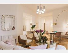 Wohnzimmer in einem modernen Apartment mit Holzboden, hellen Wänden und stilvoller Einrichtung. Es gibt eine Sitzecke mit Sofas und Sesseln, einen Glastisch mit frischen Blumen, und im Hintergrund eine offene Küche sowie einen Essbereich mit runden Tischen und Stühlen.