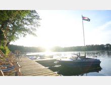Ein Holzsteg an einem ruhigen See mit mehreren Booten, darunter ein kleines Motorboot, das am Steg festgemacht ist. Links im Bild stehen Liegestühle unter einem Baum, während eine Fahne mit einem Piratenmotiv auf der rechten Seite weht; im Hintergrund ist ein bewaldetes Ufer sichtbar, beleuchtet von tiefstehender Sonne.