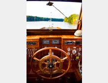 Steuerstand eines Holzboots mit einem großen, hölzernen Steuerrad, umgeben von Instrumenten und Bedienelementen. Durch das Fenster des Boots ist ein ruhiger Fluss mit bewaldetem Ufer im Hintergrund zu sehen.