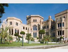 Historisches Schlossgebäude im neugotischen Stil mit sandfarbenen Fassaden, hohen Fenstern und zinnenartigen Türmen; umgeben von gepflegten Grünflächen, Topfpflanzen und dekorativen Sträuchern. Im Vordergrund ist ein angelegter Gartenweg mit Besuchern sichtbar.