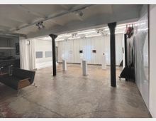 Moderner Ausstellungsraum mit Betonboden und schwarzen Metallsäulen, umgeben von weißen Vorhängen. Im Raum sind mehrere Podeste mit Skulpturen und gerahmte Kunstwerke an den Wänden zu sehen, beleuchtet durch eine Deckenkonstruktion mit integrierter Beleuchtung.