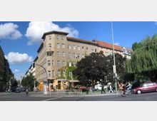 Straßenecke in einer Stadt mit einem mehrstöckigen Wohn- und Geschäftshaus aus beigefarbenem Putz, flankiert von Bäumen. Vor dem Gebäude befinden sich Geschäfte im Erdgeschoss, ein breiter Gehweg und Fußgänger sowie Radfahrer auf der Straße.